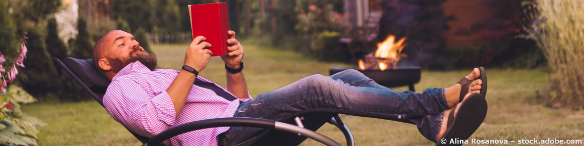 Auf dem Bild unserer Garten Relaxsessel Seite liegt ein bärtiger Mann mit pink kariertem Hemd, Jeans und Flip-Flops in einem Schaukelsessel für den Garten. Er liest in einem roten Buch, das er in den Händen hält und sieht sehr entspannt aus.